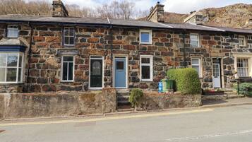 A stone building with multiple doors and windows, a small garden area with a bench, and a clear road in front.