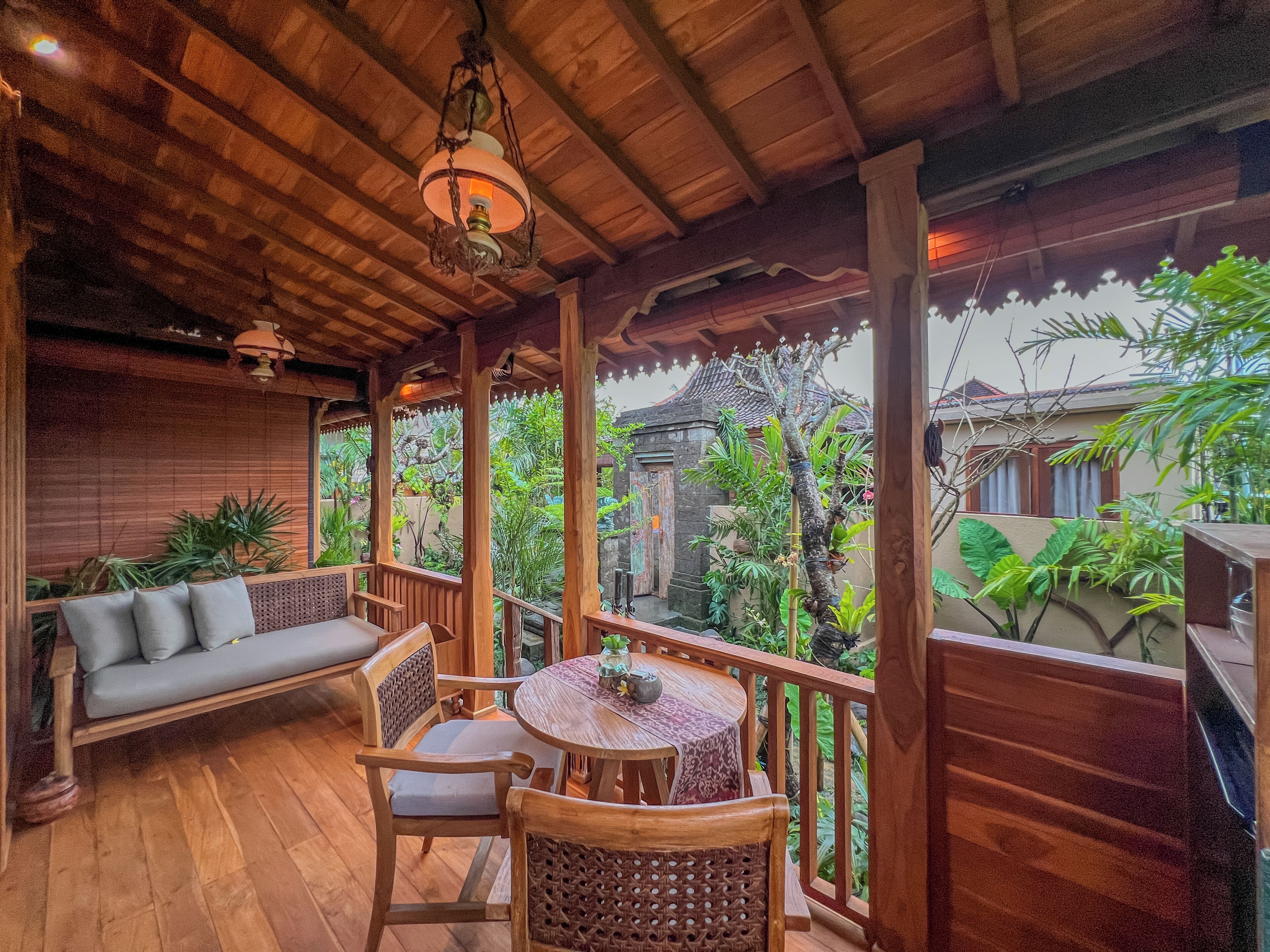 Una terraza de madera con un sofá y sillones, rodeada de exuberante vegetación y un edificio tradicional al fondo.