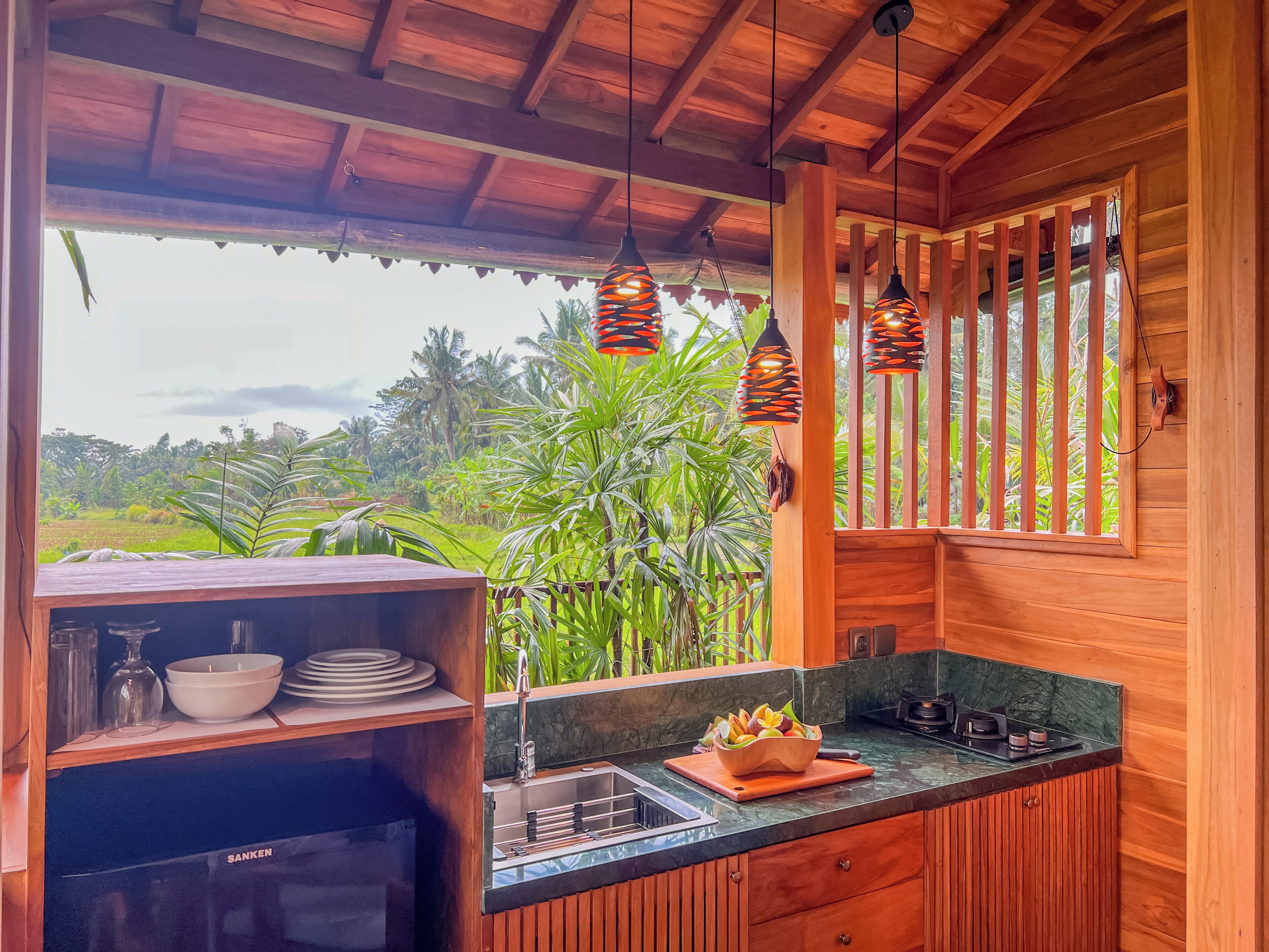 Interior de madera con cocina, un lavamanos y ventana con vista a un jardín exuberante.