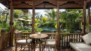 A wooden deck with a round table and chairs, a sofa, and a view of a pool and tropical vegetation. - Edge of Ubud by GenuineHost (Ubud)