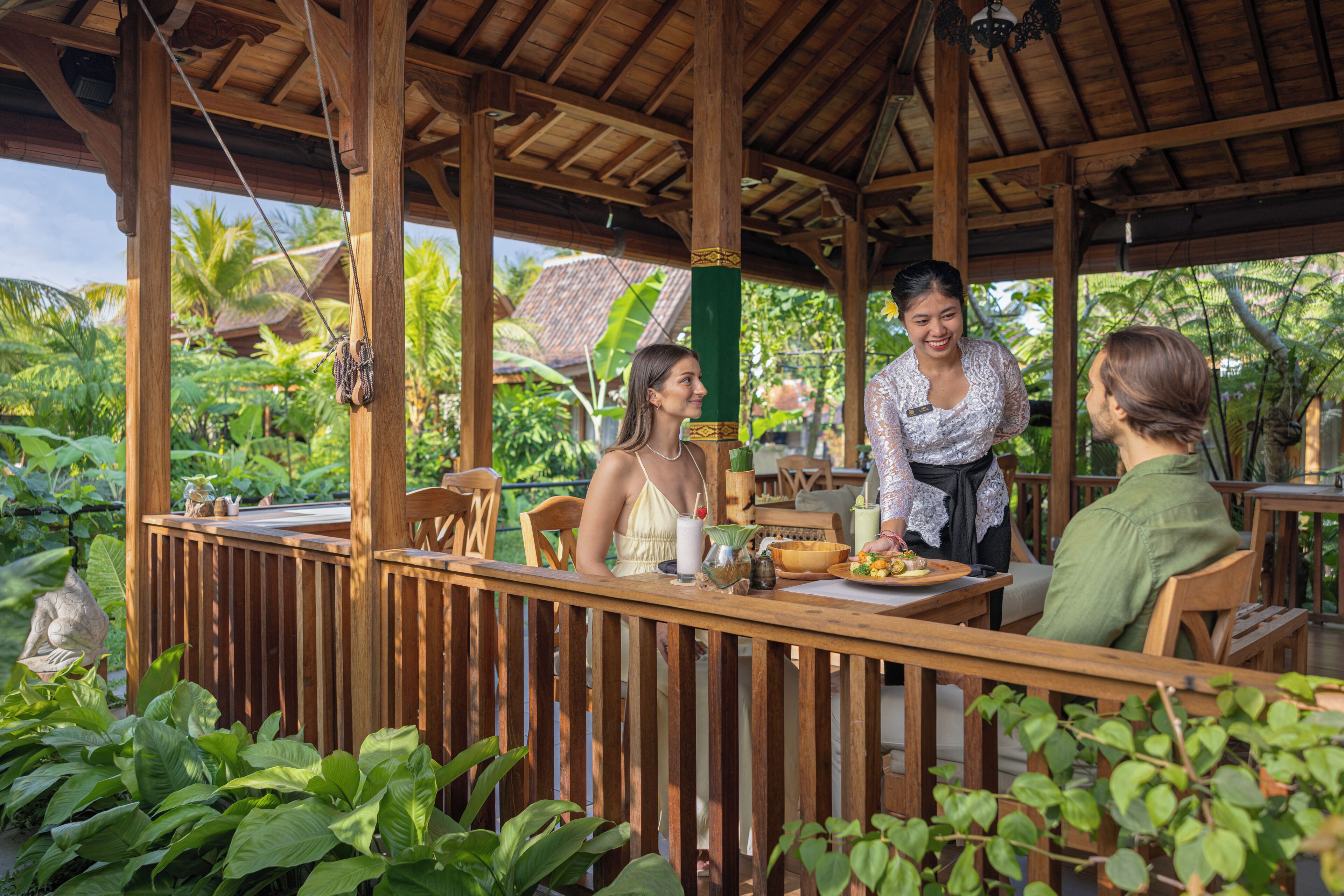 A wooden outdoor dining area with a thatched roof, surrounded by lush greenery and tropical plants.