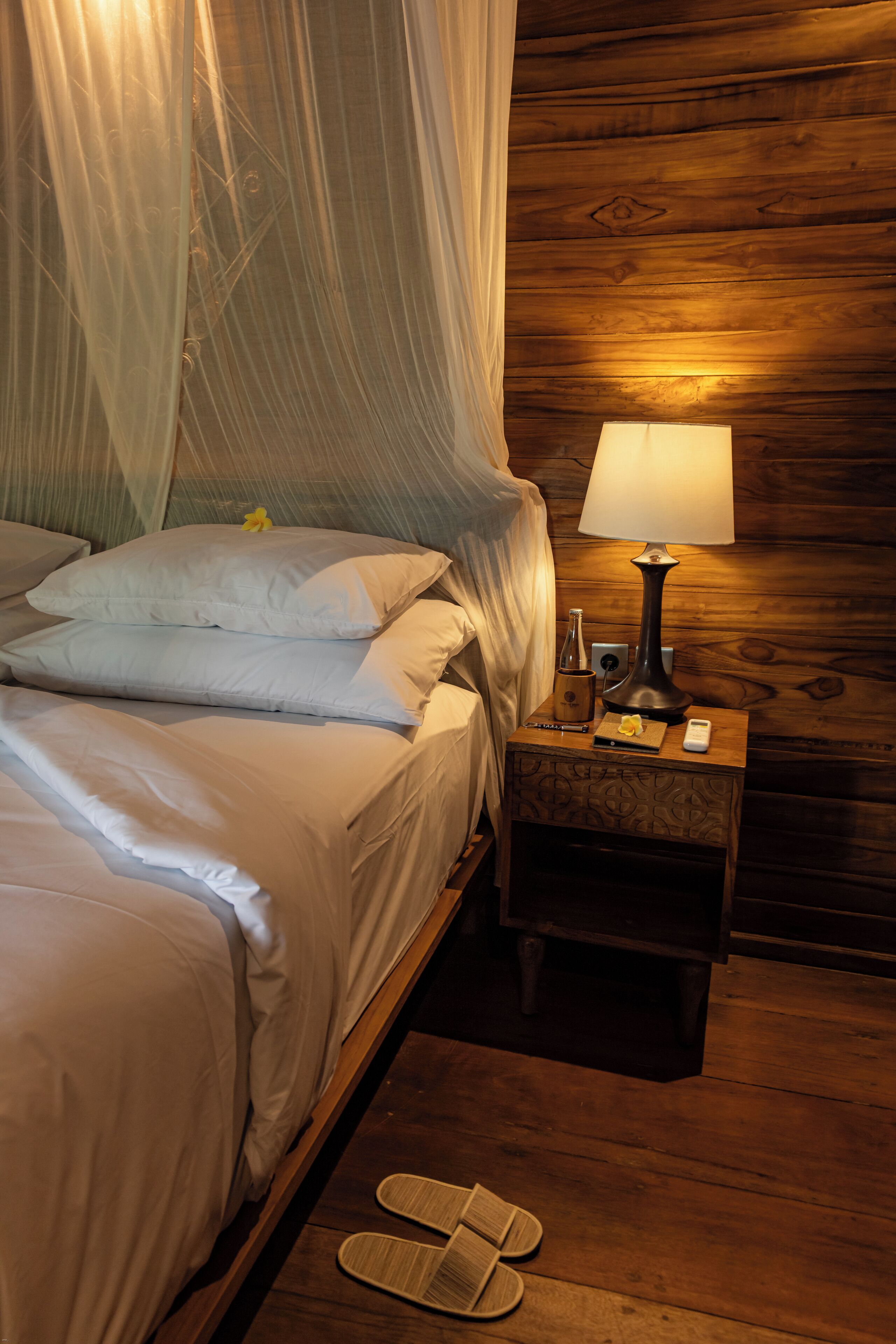 A neatly made bed with white linens and pillows, a bedside table with a lamp, and a pair of slippers on the floor.
