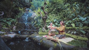 Una mujer vestida con traje tradicional sentada sobre una roca junto a una cascada en un bosque frondoso y verde.