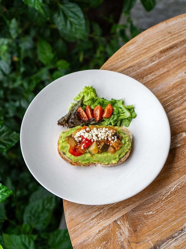 A plate with a sandwich on a white plate, placed on a wooden table with a green leafy background. - DITUJU Ubud by GenuineHost (Sukawati)