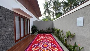Un chemin bordé d’un paillasson et d’un tapis décoratif à motifs floraux qui mène à un bâtiment doté d’un mur en pierre et d’un plancher en bois.