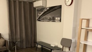 Un salon moderne avec une table basse en verre, des fauteuils gris et marron, un canapé, une petite table d’appoint, une horloge au mur et une vue sur le paysage urbain par une fenêtre.