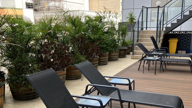 Des fauteuils de salon d’extérieur sont disposés sur une terrasse en bois, agrémentée de plantes en pot, avec un bâtiment visible à l’arrière-plan.