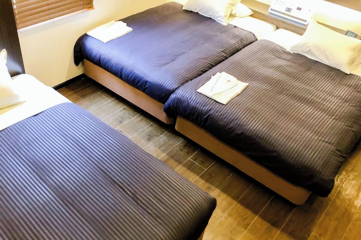 A hotel room with two single beds, each with a pillow and a folded towel. The room has wooden flooring and a window with blinds.