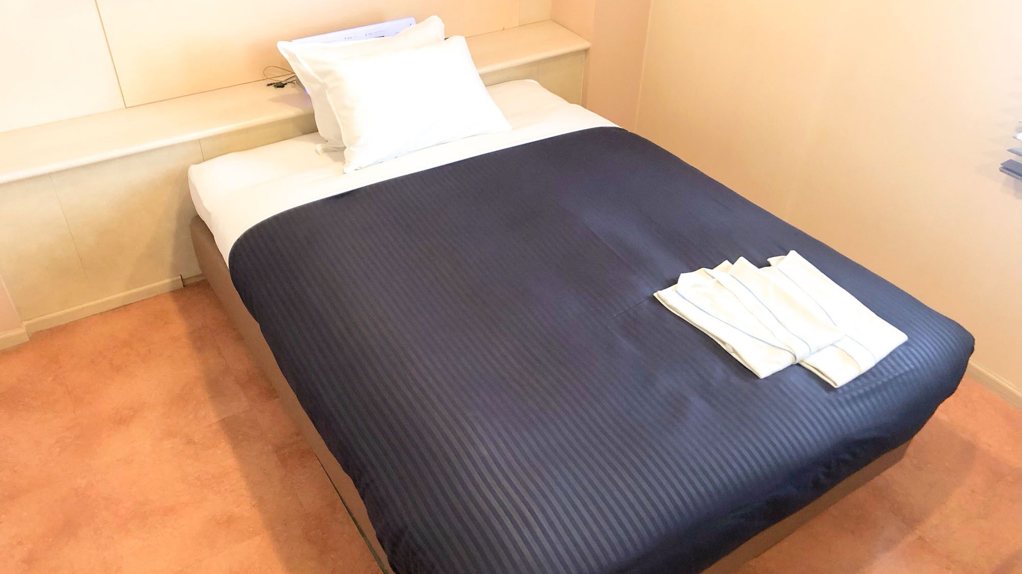 Een eenpersoonsbed met een donkerblauwe sprei en witte kussens, een klein nachtkastje met een lamp, en een vloerbedekking.