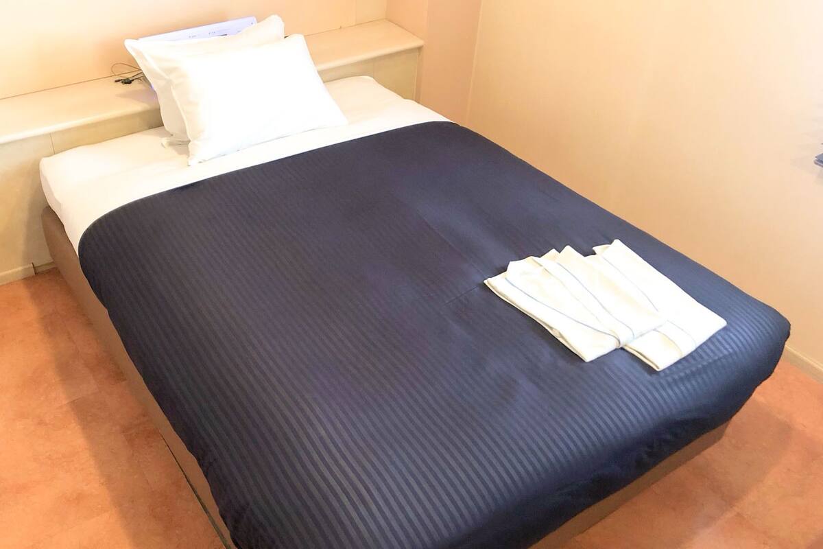A single bed with a dark blue bedspread and white pillows, a small bedside table with a lamp, and a carpeted floor.