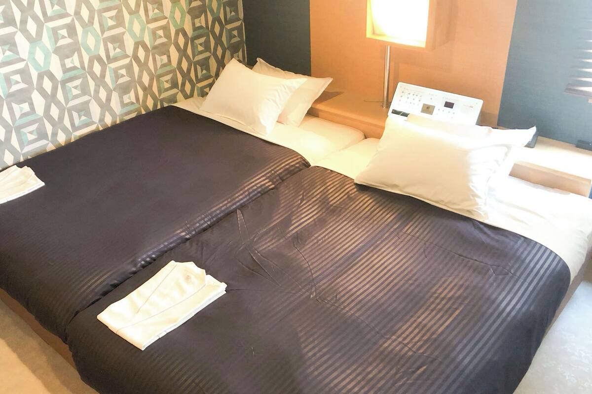 A hotel room with a bed, pillows, a bedside table, and a lamp.