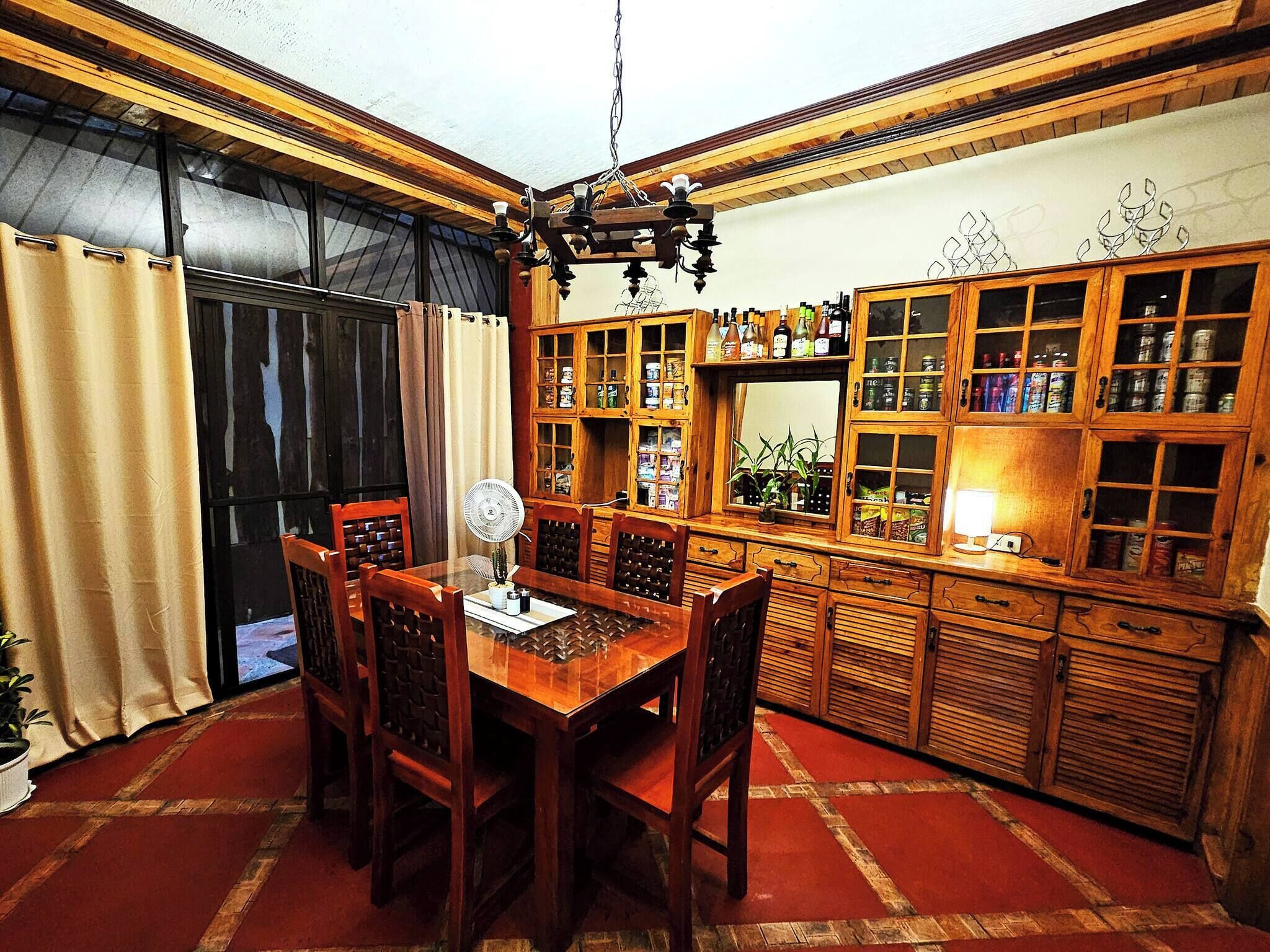 A traditional wooden dining area with a table and chairs, a fan, and a kitchen area with wooden cabinets and shelves.