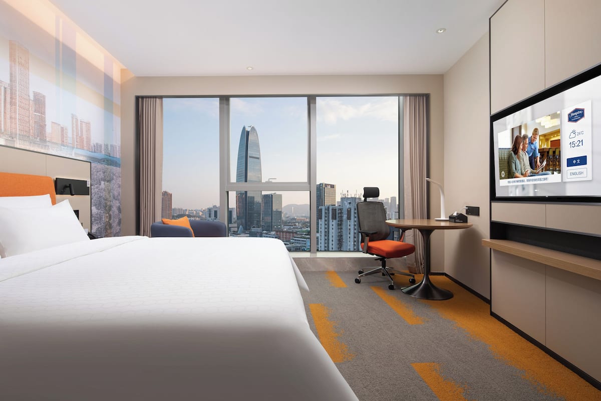 Habitación de hotel con una cama grande, un escritorio, una silla y un televisor que muestra una vista urbana.