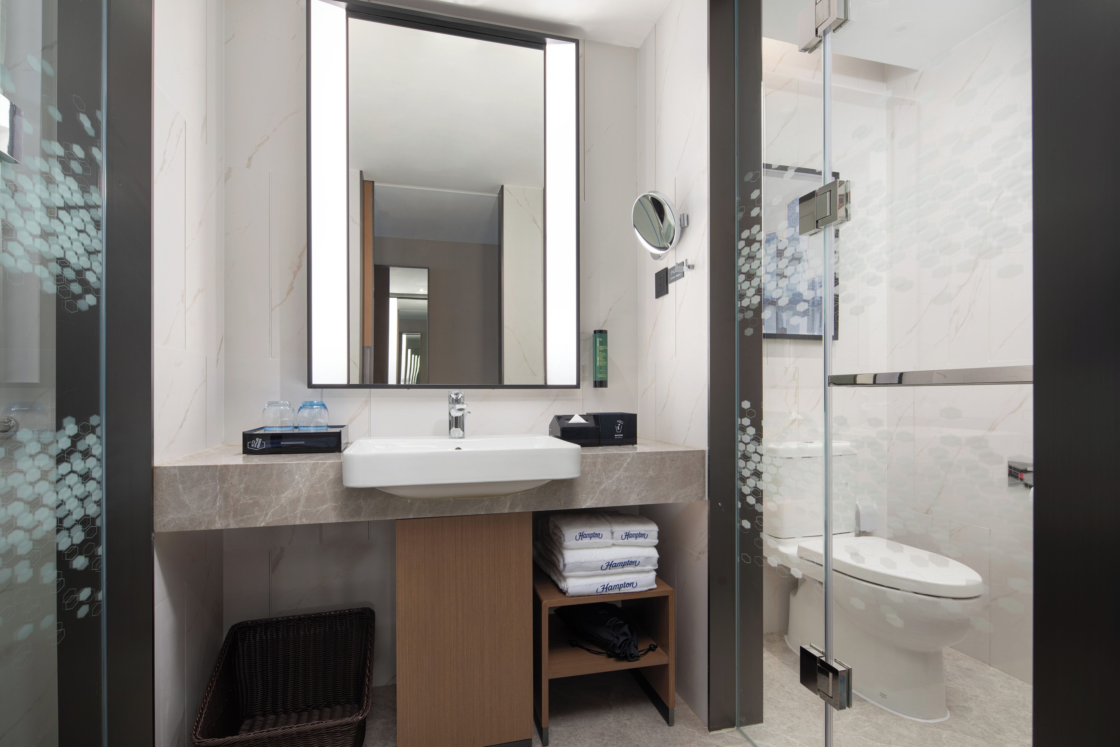 A modern bathroom with a large mirror, a sink, and a toilet. There is a wicker basket and a shelf with towels.