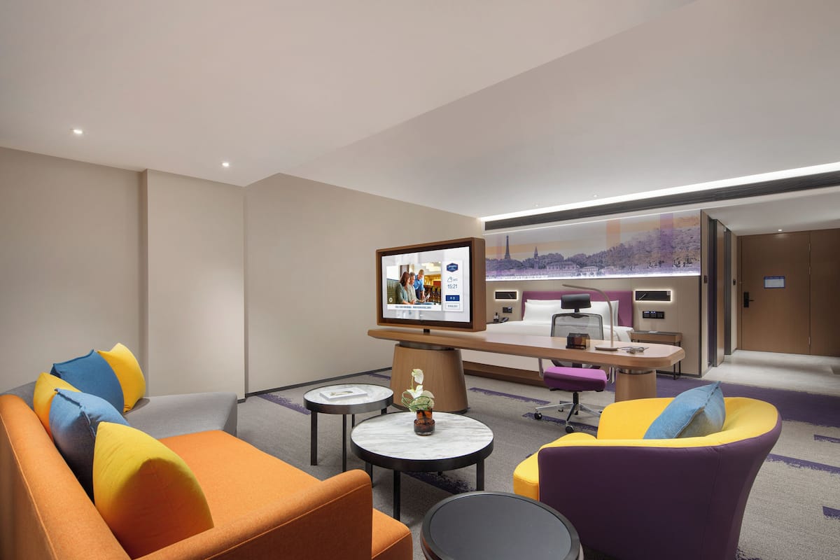Una habitación de hotel moderna con sofá, sillones, una mesita, televisor y escritorio con computadora.