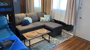 A cozy room with a sectional sofa, a small table with woven placemats, a bunk bed, and a large window with curtains. - Adorable 1-bedroom condo with kitchen, pool, WiFi near Waikiki Beach, sleeps 6-8 (Honolulu)