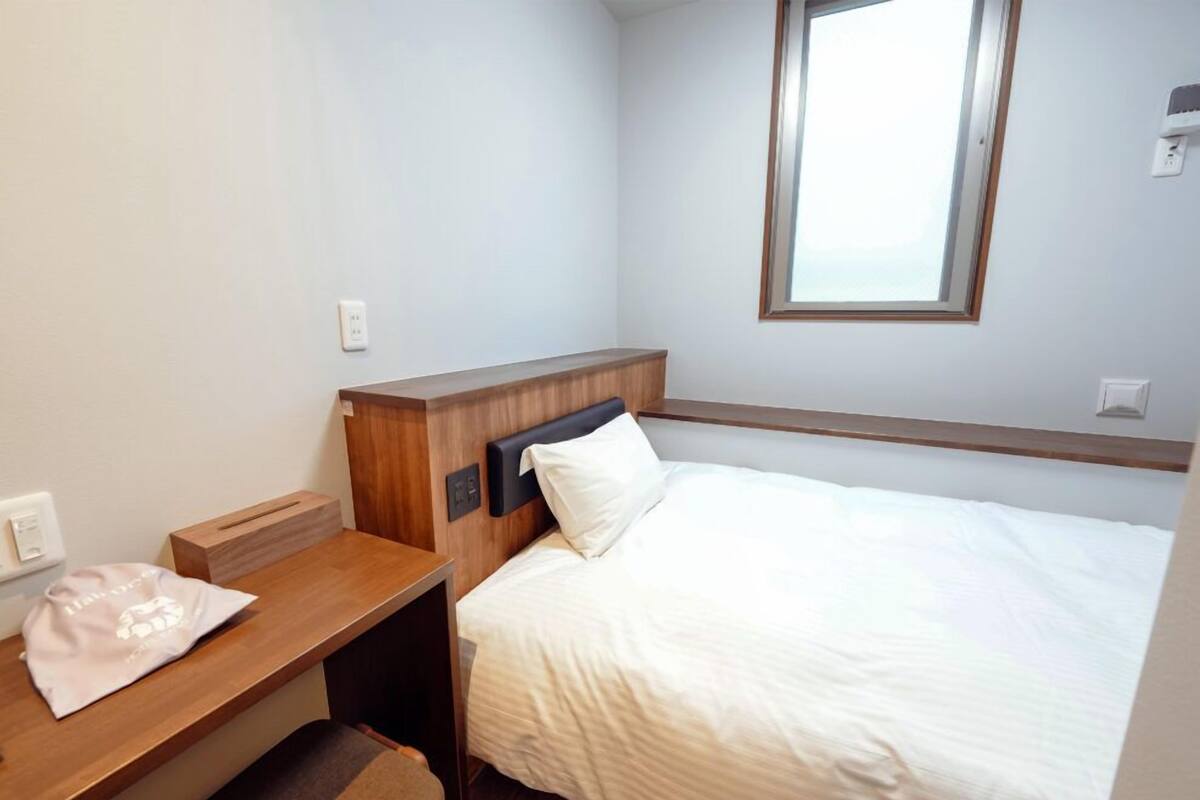Una habitación de hotel con una cama, una mesita de noche de madera, una pequeña mesa y una ventana.