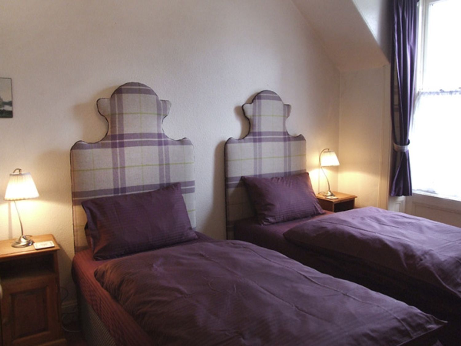 A room with two beds, each with a headboard and a matching pillow. There are bedside tables with lamps and a window with curtains.