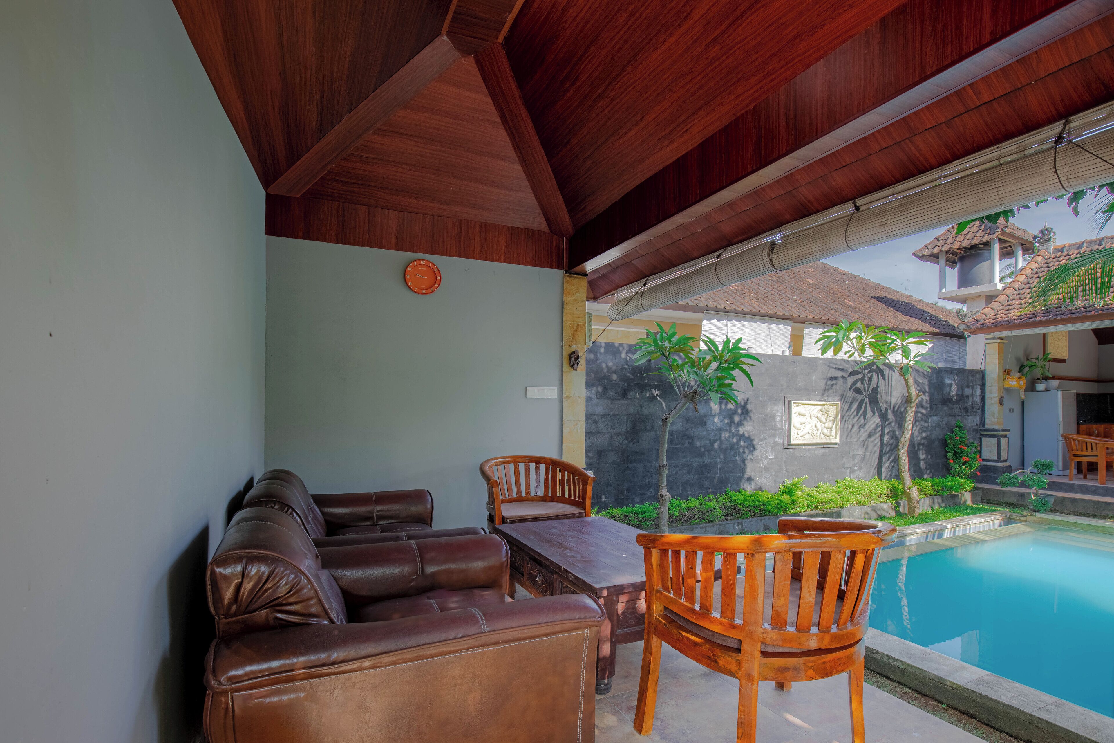 A covered outdoor seating area with a pool, wooden furniture, and a stone wall.
