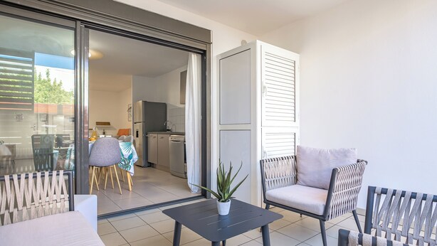 Une porte-fenêtre coulissante donne sur une cuisine et un coin repas, où se trouvent une table et des chaises, une plante en pot posée sur la table et un canapé.