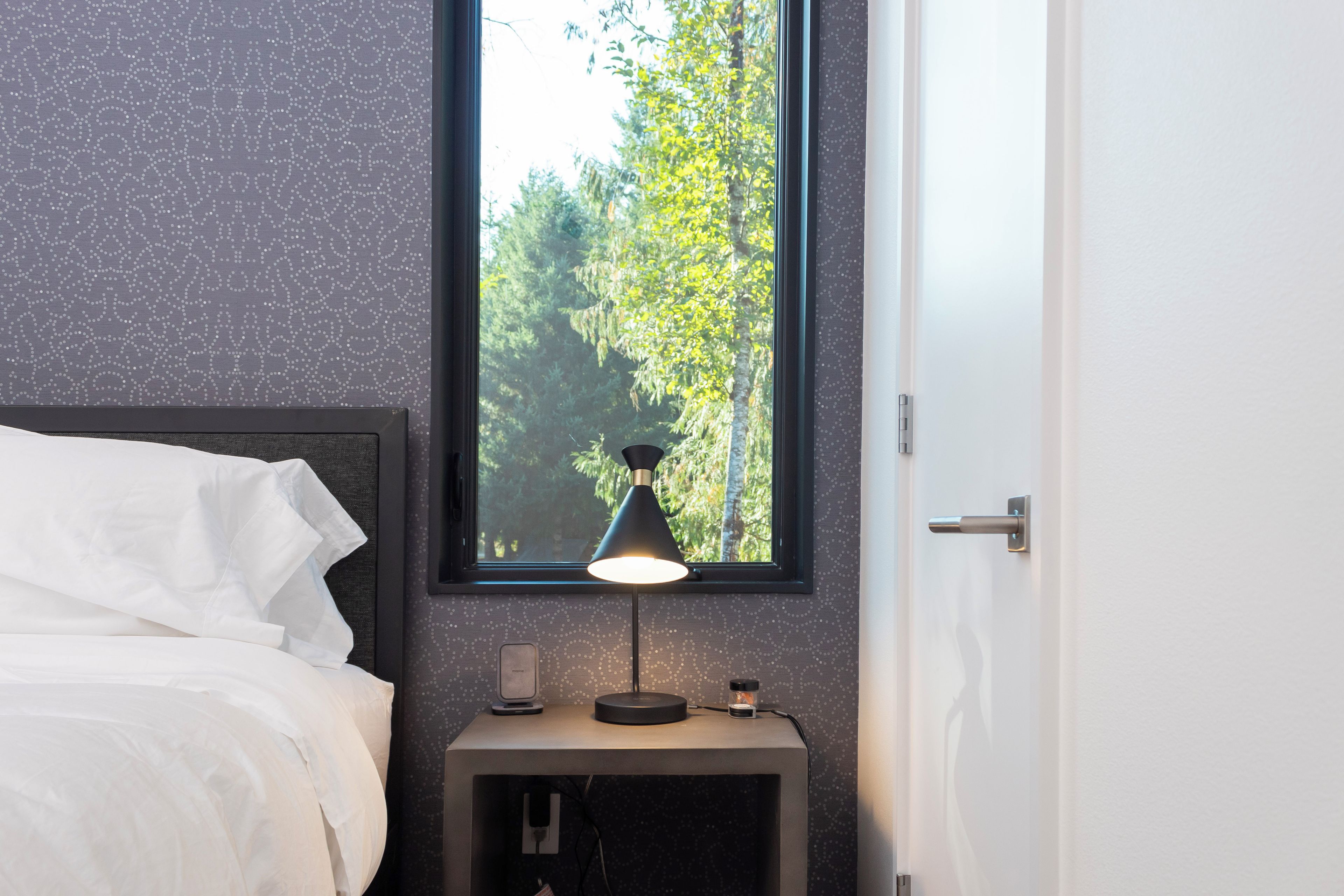 A neatly made bed with white linens, a bedside table with a lamp, a small clock, and a window with a view of trees.