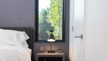 A neatly made bed with white linens, a bedside table with a lamp, a small clock, and a window with a view of trees.