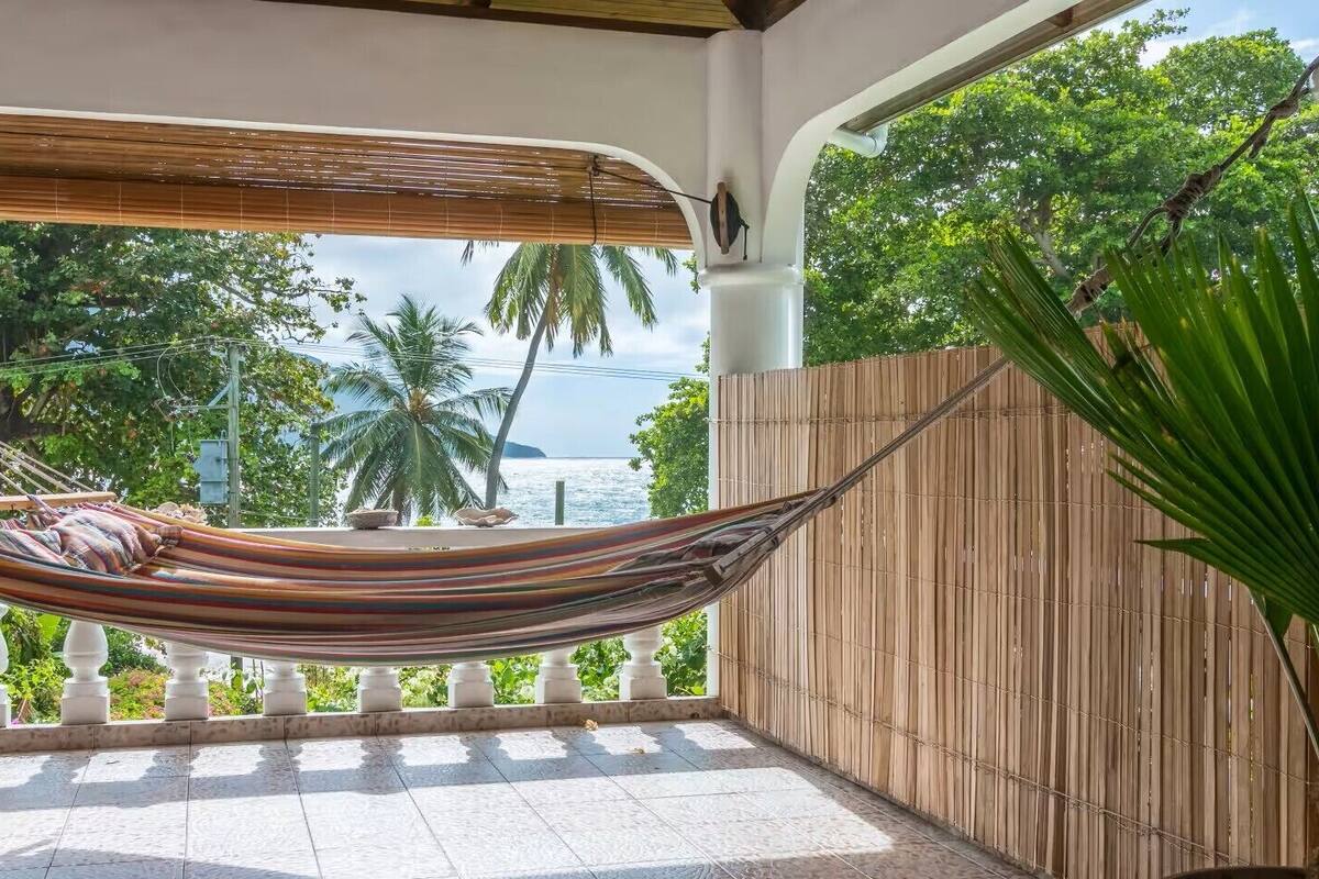 A hammock on a balcony with a view of the sea and palm trees.