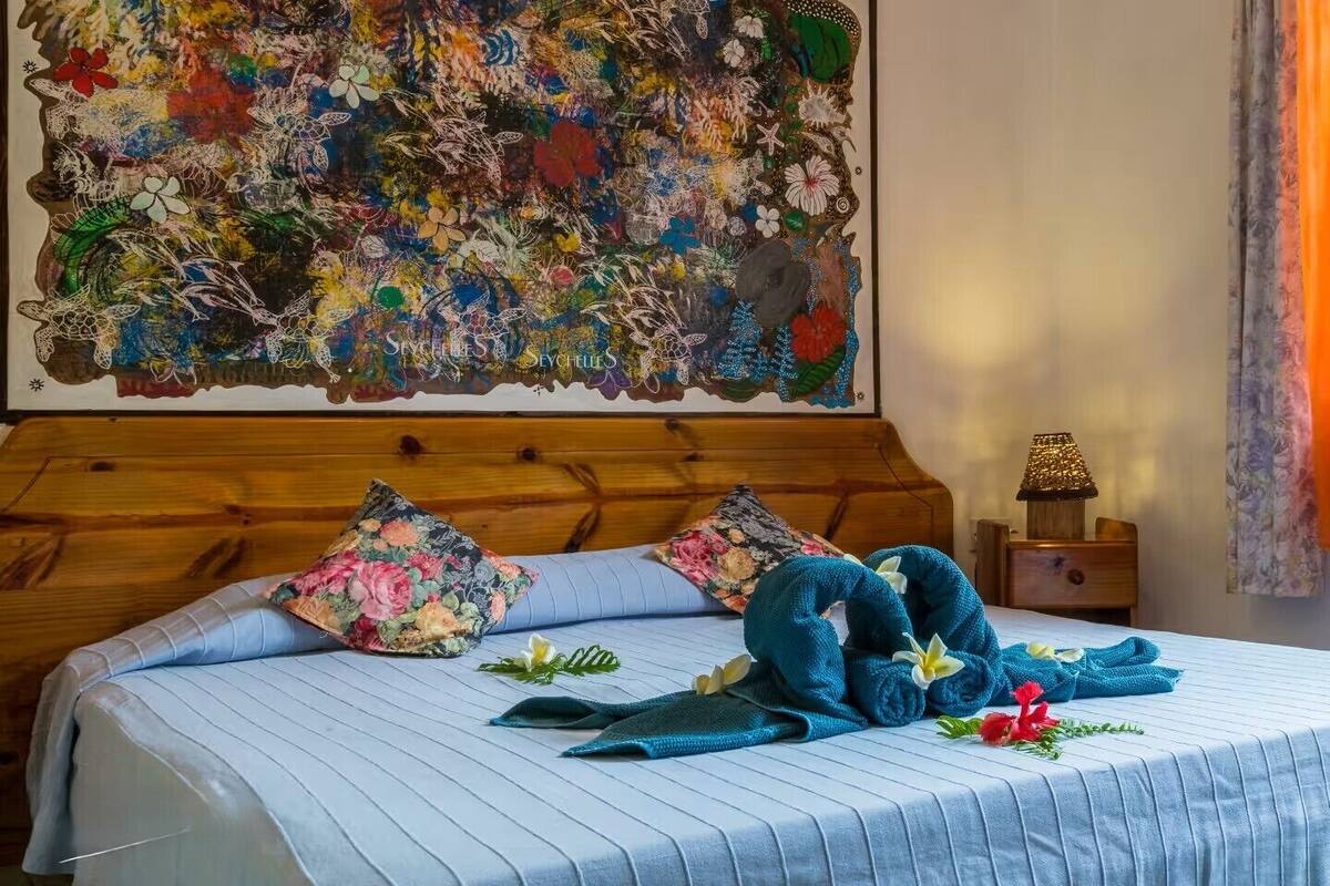 A neatly made bed with a blue bedspread and floral pillows, a wooden headboard, a nightstand with a lamp, and a large floral artwork on the wall.