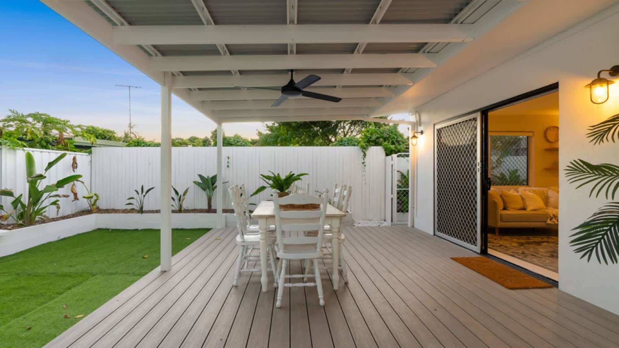 A covered patio with a table and chairs, a ceiling fan, and a white fence in the background.