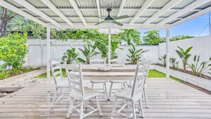 Eine überdachte Terrasse im Freien mit Tisch und Stühlen, umgeben von einem weißen Zaun und üppigem Grün.