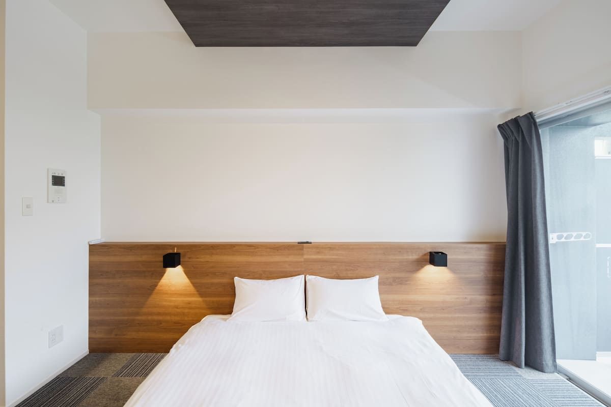A modern hotel room with a wooden headboard, a bed with white linens, two wall-mounted lights, and a large window with curtains.