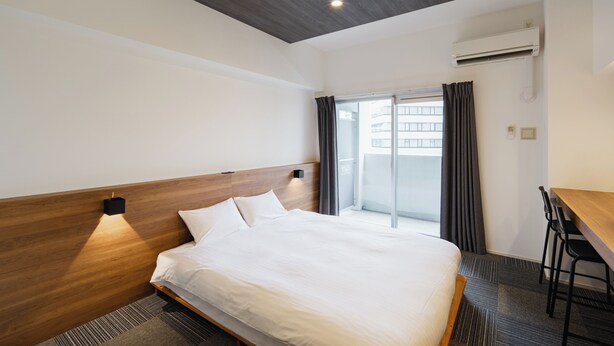 A hotel room with a wooden headboard, a bed with white bedding, a desk with two chairs, and an air conditioning unit. - HIT Tenjin (Fukuoka)