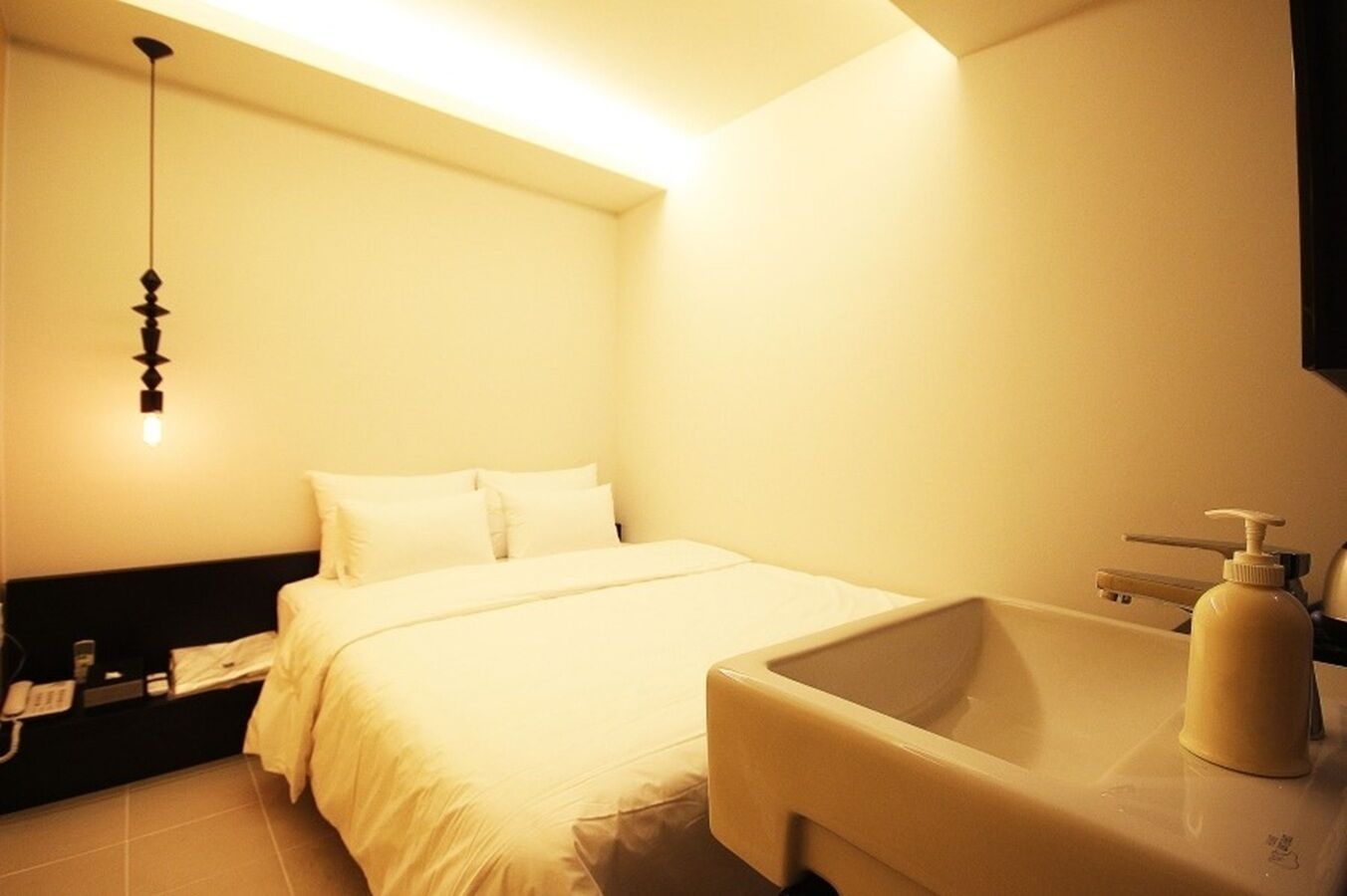 A hotel room with a bed, a bedside table, a nightstand, a wall-mounted light fixture, and a bathroom with a bathtub and a soap dispenser.
