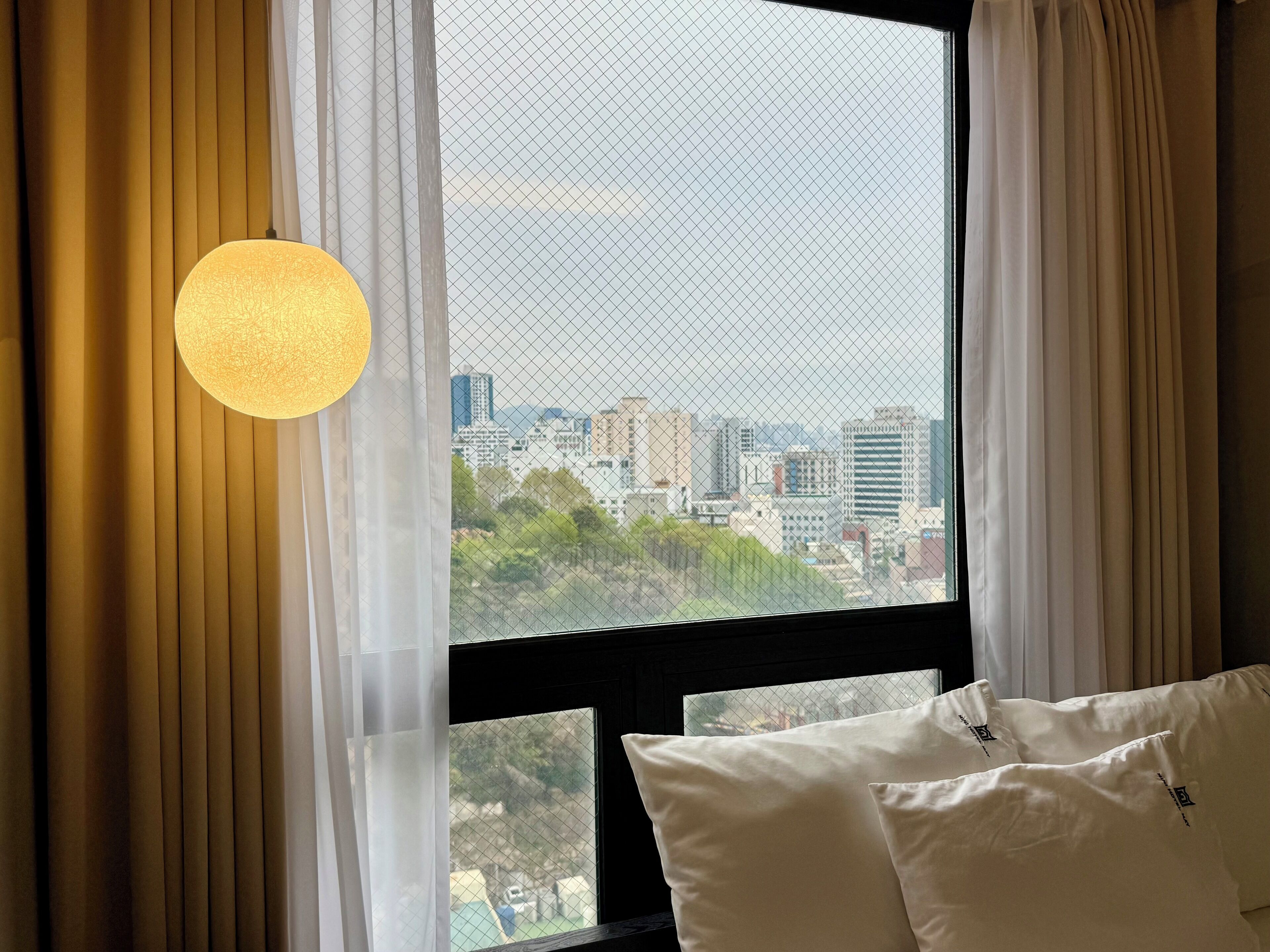 Habitación de hotel con un ventanal grande, cortinas blancas y una lámpara redonda.