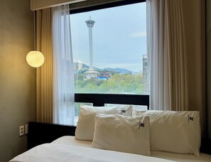 Habitación de hotel con cama, almohadas blancas y ventana con vistas a una torre y vegetación.