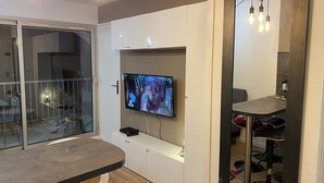 A modern living room with a flat-screen TV mounted on a white cabinet, a dining table with chairs, and a large mirror with hanging lights.
