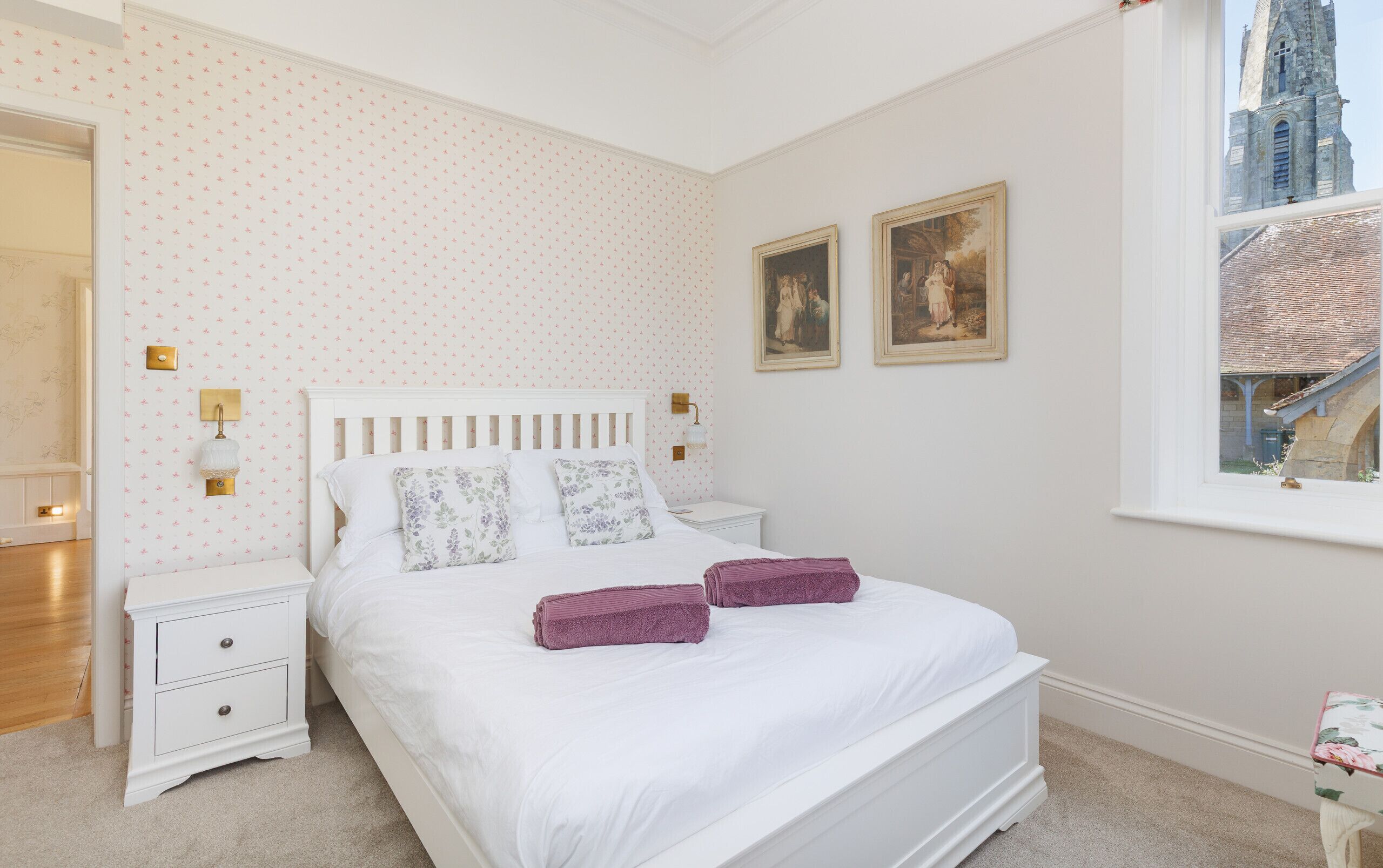 A bedroom with a white bed, two nightstands, a window with a view of a building, and two framed pictures on the wall.