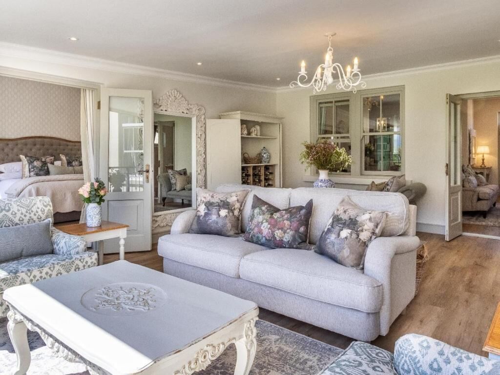 A spacious living room with a large sofa, a coffee table, and a chandelier. There is a bed with a headboard and a window with curtains.