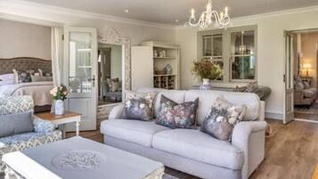 A spacious living room with a large sofa, a coffee table, and a chandelier. There is a bed with a headboard and a window with curtains.
