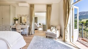 Ein Schlafzimmer mit Bett, Schreibtisch, Stuhl, Schminktisch mit Spiegel und Blick auf einen Balkon mit schöner Landschaft.