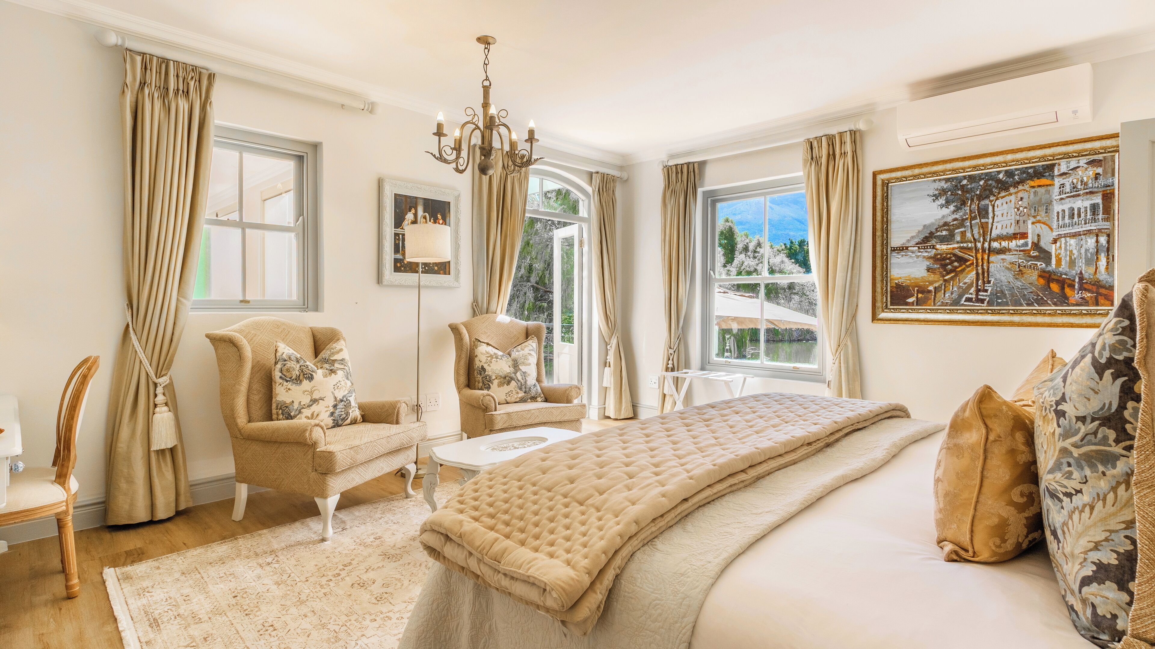 A spacious bedroom with a large bed, two armchairs, a floor lamp, and a painting on the wall.