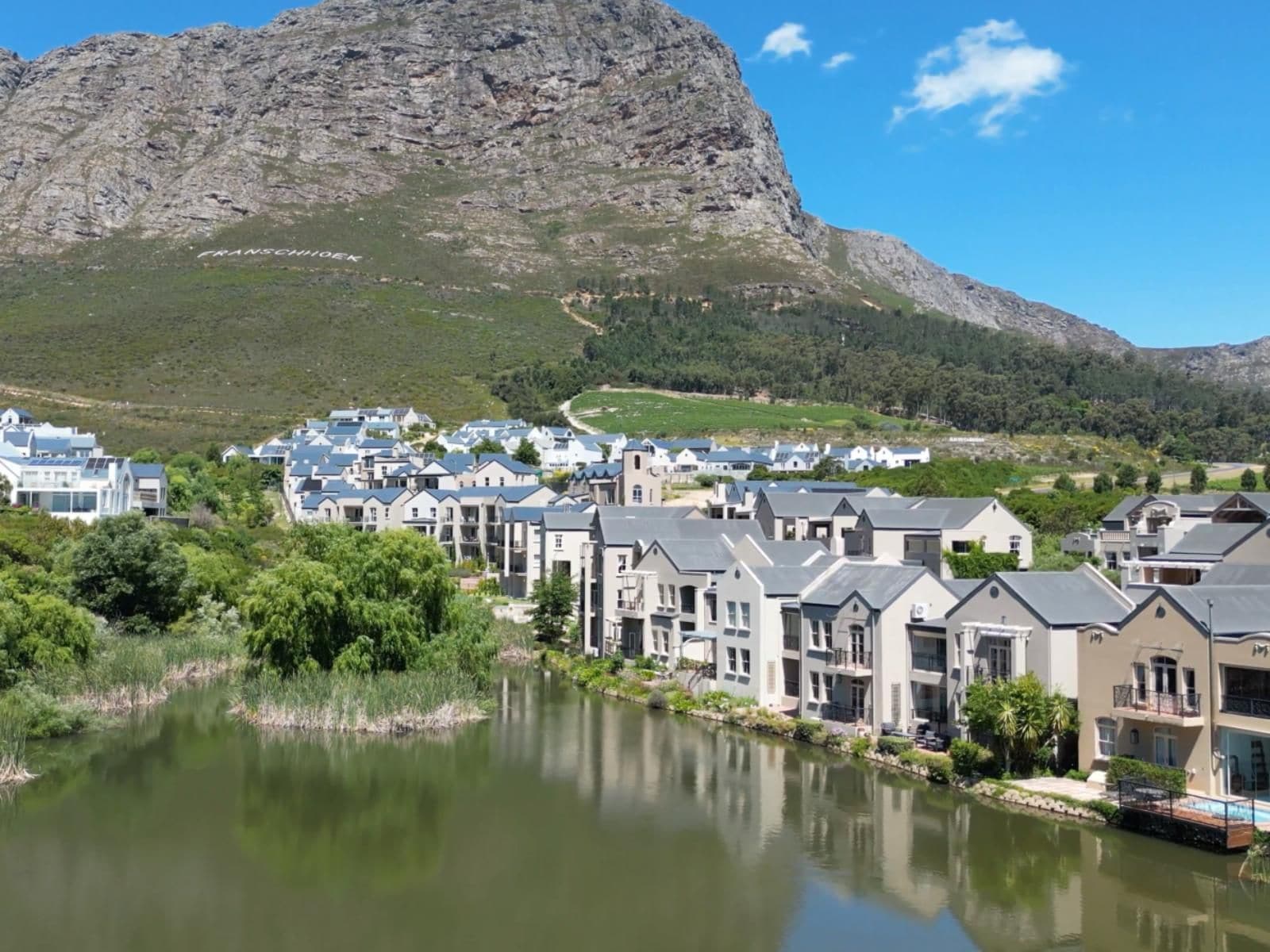 Un quartier résidentiel avec des maisons le long d’une rivière, une montagne en arrière-plan et un ciel dégagé.