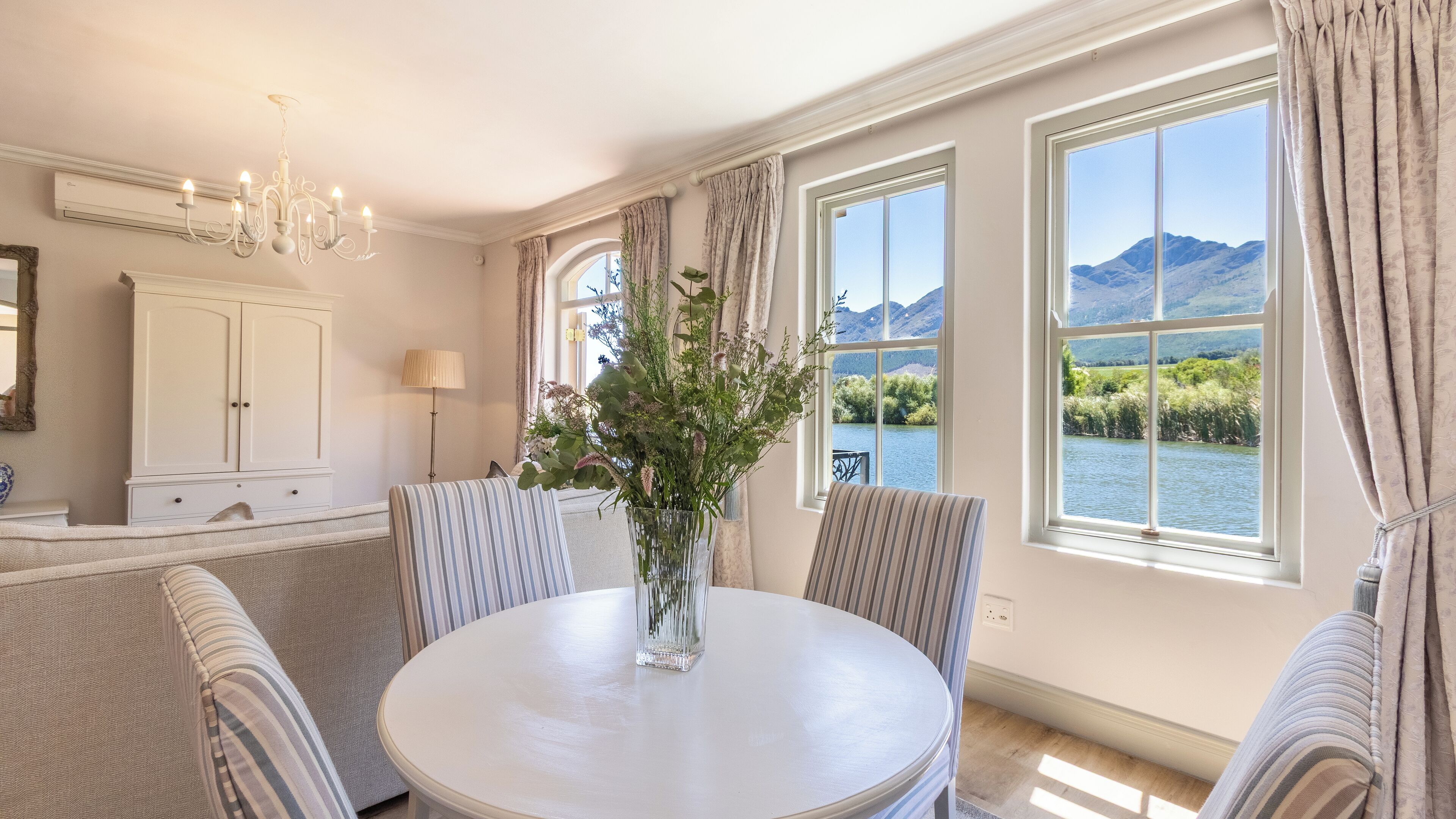 A dining area with a round table, striped chairs, a vase with flowers, and a view of mountains and water through large windows.
