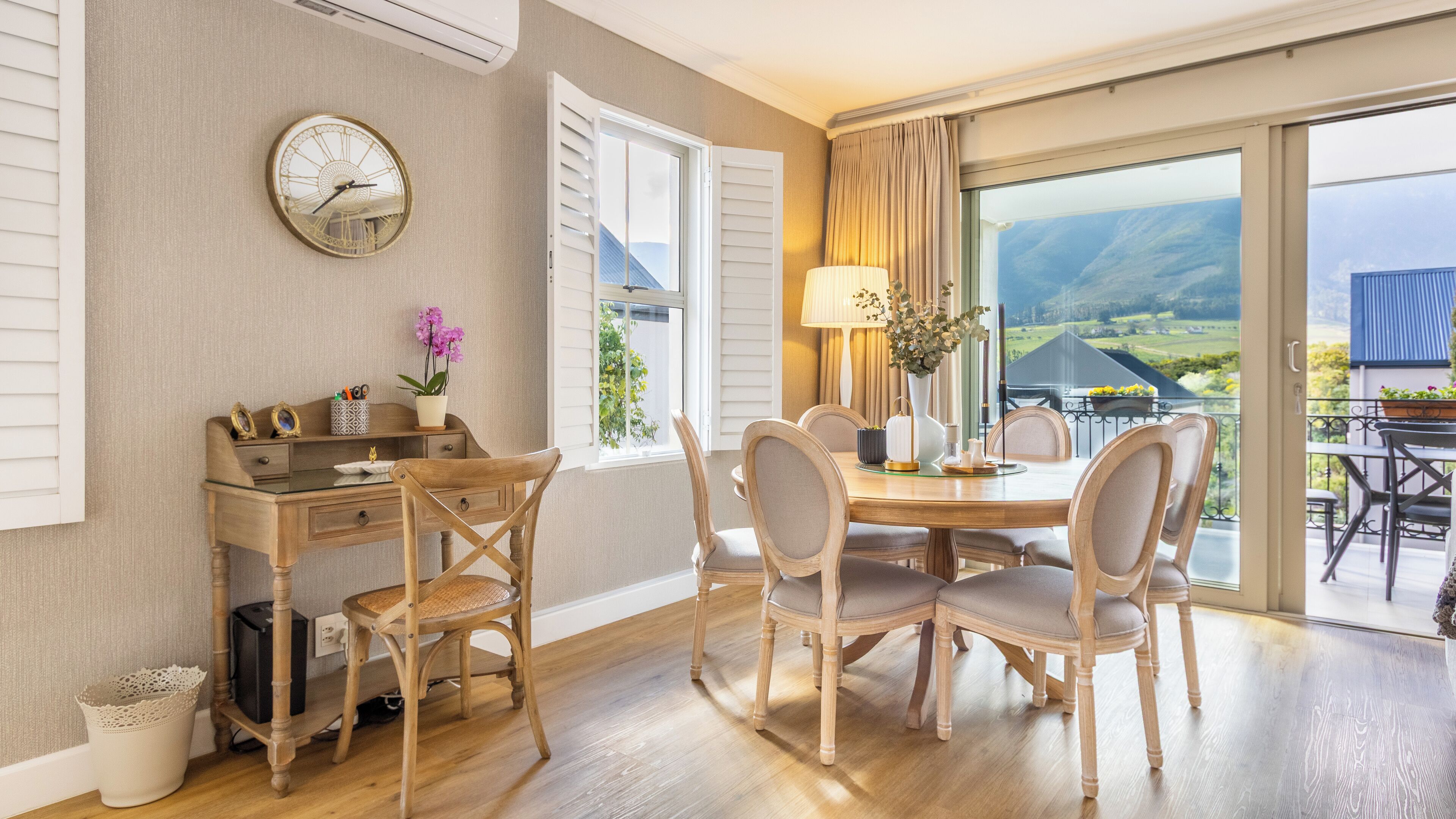A dining area with a wooden table and chairs, a clock on the wall, and a view of the outdoors.