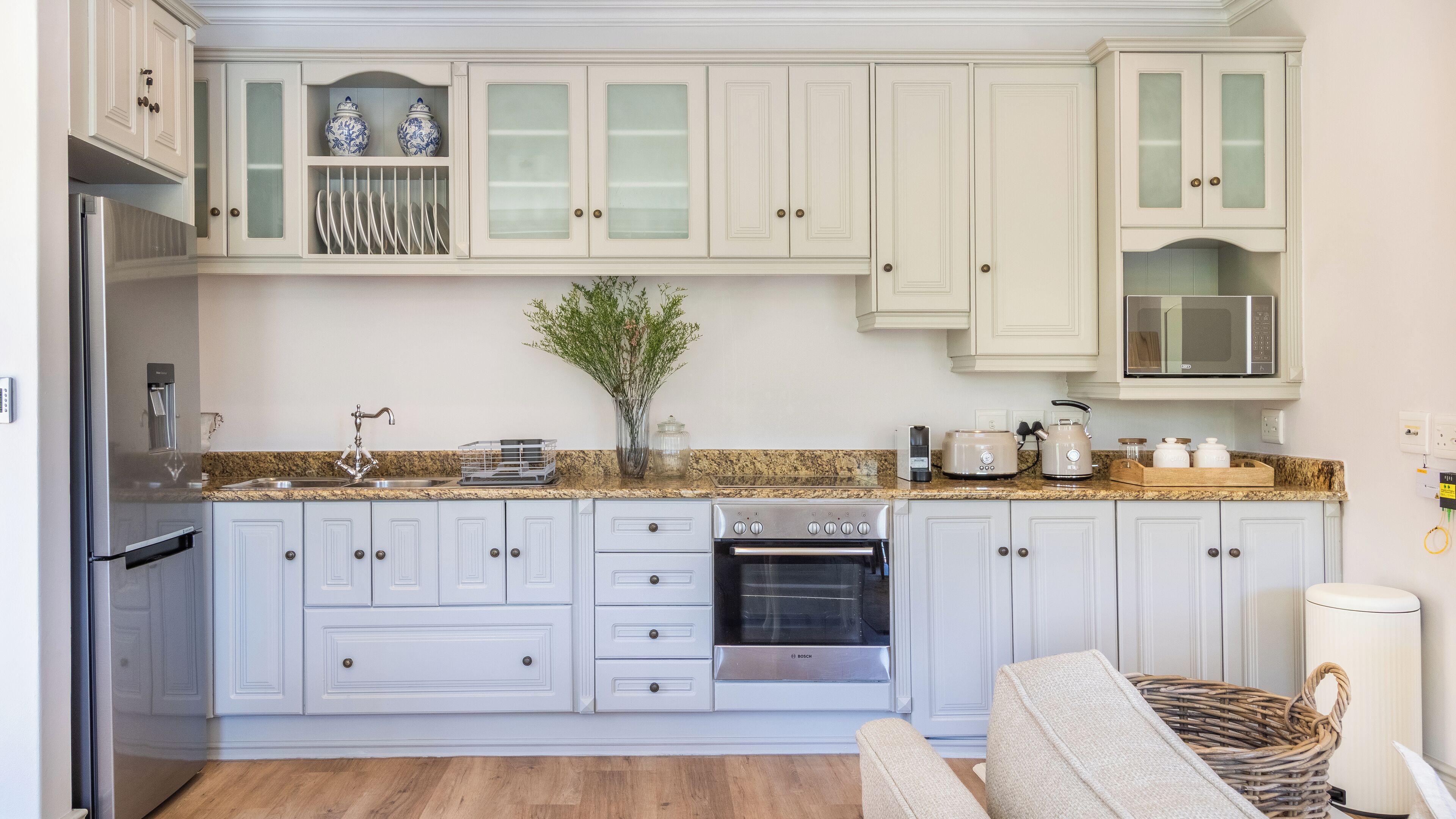 Una cocina con armarios blancos, horno Bosch, microondas y refrigerador de acero inoxidable.