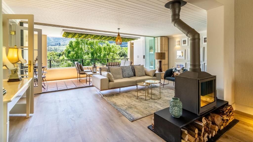 A modern living room with a sofa, coffee table, and a fireplace. There is a balcony with a view of greenery and mountains.