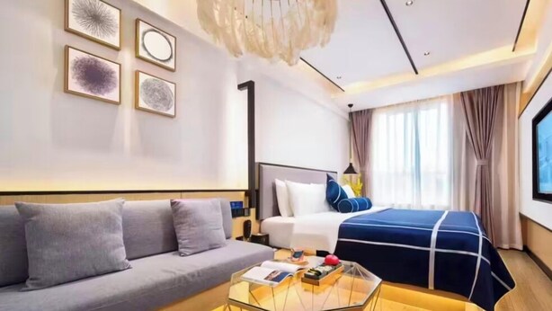 A modern hotel room with a sofa, a bed with a blue cover, a glass coffee table, and artwork on the wall. - Z Holiday Hotel (Chengdu)