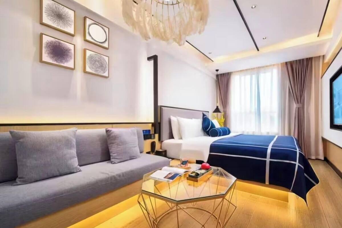 A modern hotel room with a sofa, a bed with a blue cover, a glass coffee table, and artwork on the wall.