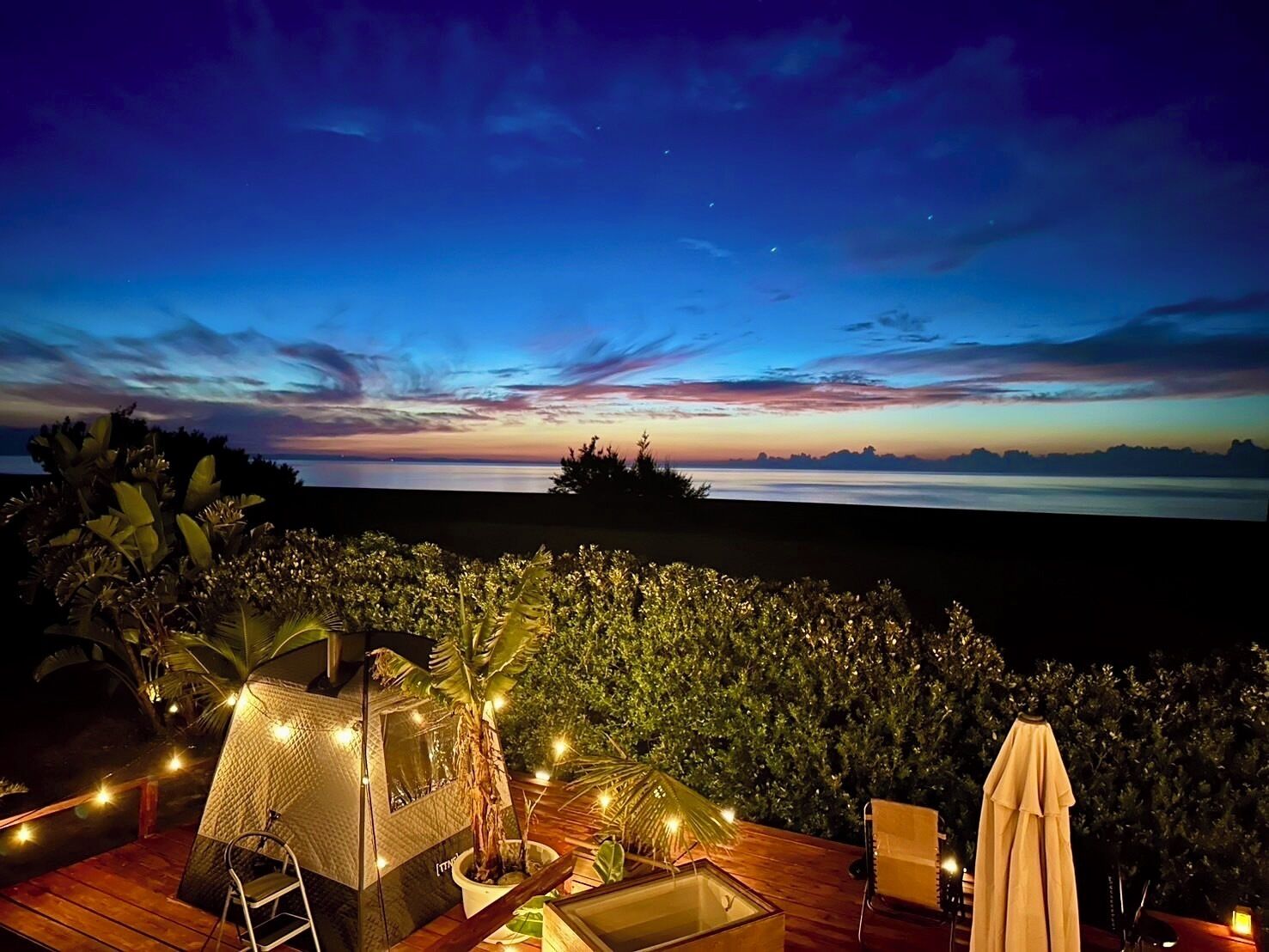 A deck with a tent, chairs, and a table, overlooking a body of water at dusk.