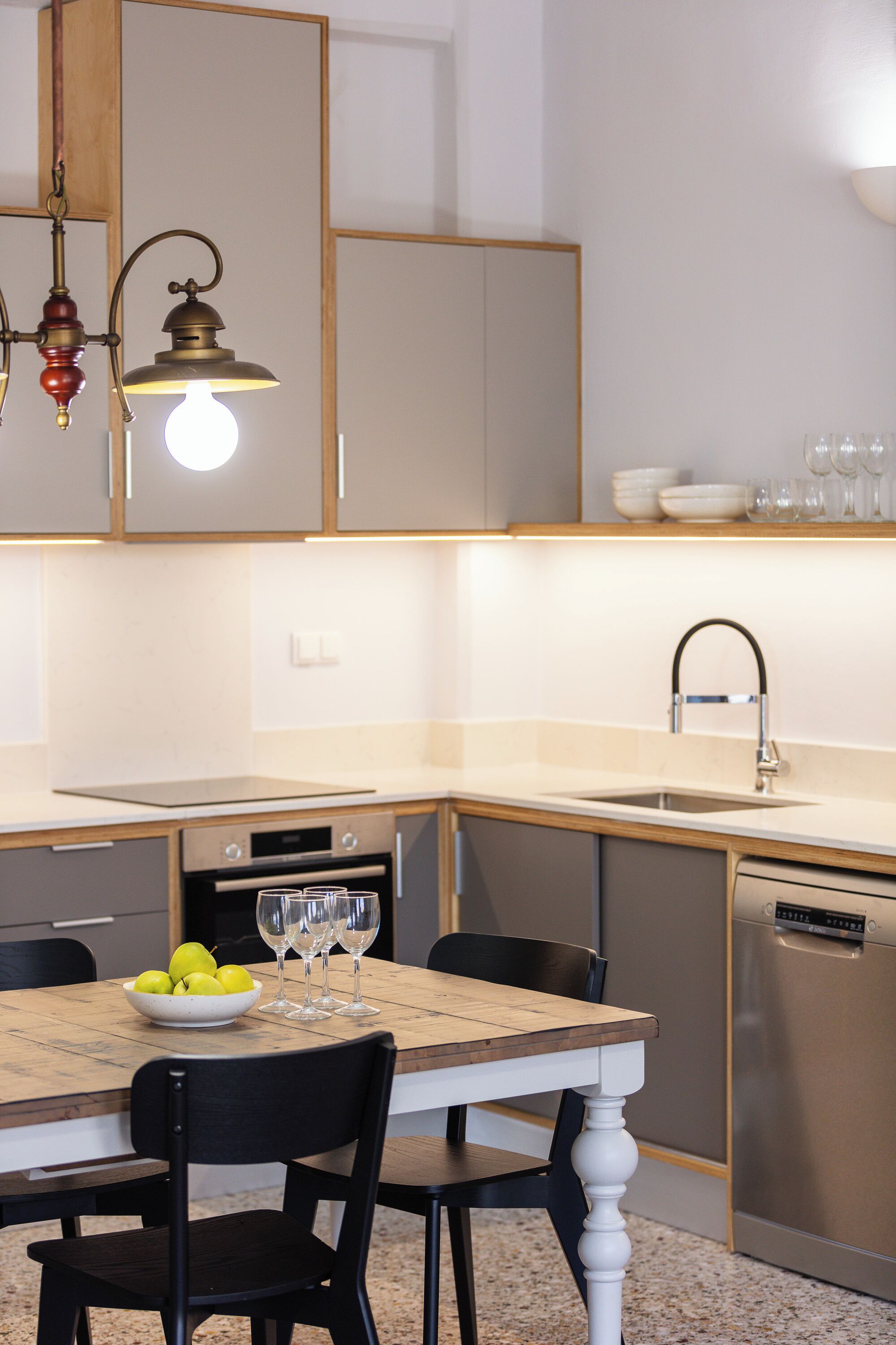 Una cocina moderna con una mesa de madera, sillas negras, un cuenco de fruta y copas de vino.
